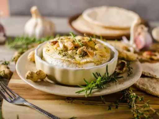 Pieczony camembert w naczyniu, ozdobiony ziołami i orzechami, z czosnkiem i pieczywem w tle.