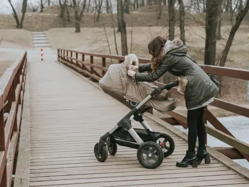 Kobieta na drewnianym moście poprawia gondolę wózka. Zastanawiasz się, jaki wózek wybrać mamaginekolog? Ten model wygląda na solidny.