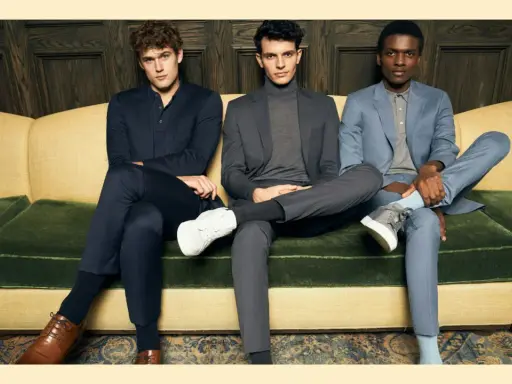 Three men in suits sit on a couch. The man on the left wears dark dress pants, while the other two wear suit pants.