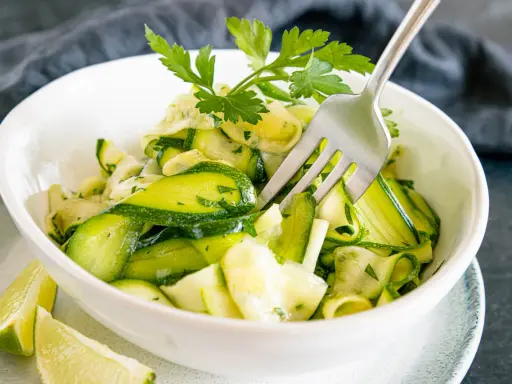 Frischer Zucchini-Salat mit Petersilie und Limettenspalten, serviert in einer weißen Schüssel. Eine Gabel steckt im Salat.