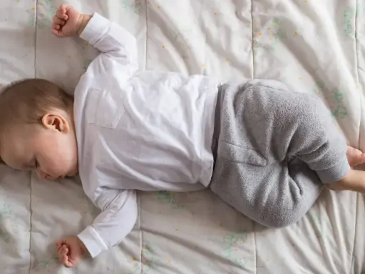 Un neonato non dorme, ma giace sdraiato su una coperta con un pigiamino bianco e grigio.