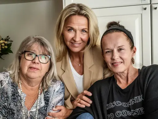 Tre kvinnor, möjligen lill babs barn, ler mot kameran. En kvinna i mitten bär en beige kavaj, den vänstra har glasögon och den högra en svart tröja.