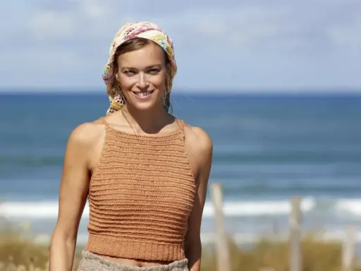 Woman in a crocheted top and headscarf smiles at the camera on a beach.