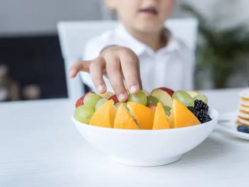 Małe rączki sięgają po miskę pełną owoców: pomarańczy, truskawek, jabłek i winogron dla niemowlaka.
