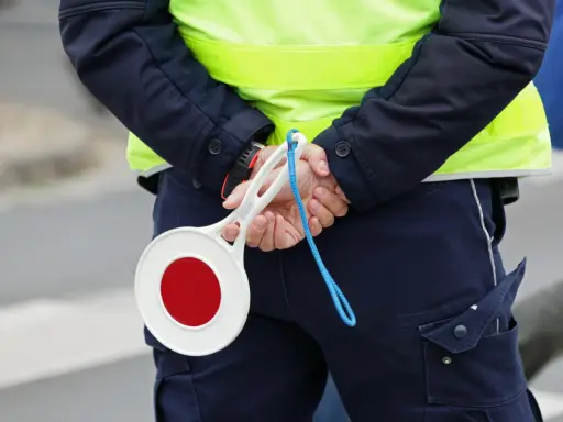 Policjant w kamizelce odblaskowej trzyma za plecami lizak do zatrzymywania pojazdów.