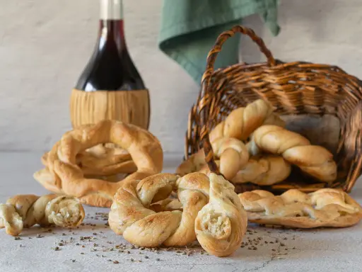 Pane intrecciato e ciambelle, ispirazione per ricette ciociare, accanto a una bottiglia di vino rosso e un cesto di vimini.
