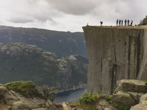Grupa ludzi na krawędzi klifu w **Stavanger, Norwegia**, z widokiem na fiord i góry.