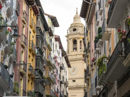 Edificio con balcónes floridos y torre de iglesia en Navarra. ¿Buscas ayudas cambio ventanas Navarra?