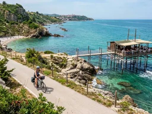 Ciclista lungo la via dei trabocchi, con un trabocco in legno sull'acqua cristallina e una spiaggia rocciosa.