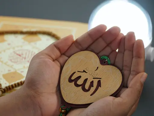 Hände halten ein Herz mit dem Wort "Allah" in arabischer Schrift, ein Symbol für ikhlas sure. Gebetskette und ein Buch liegen im Hintergrund.