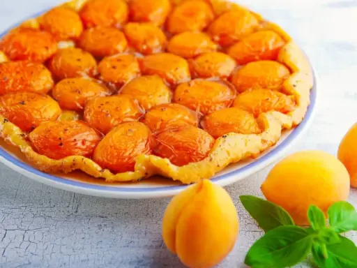 Tarte aux abricots caramélisés sur un fond de tarte croustillant, avec des abricots frais et des feuilles de basilic sur le côté.