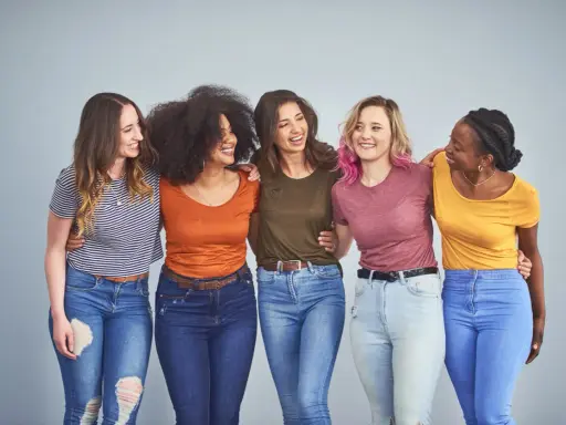 Cinque amiche sorridono, vestite con jeans e magliette colorate, tra cui spiccano tonalità marrone e bianco.