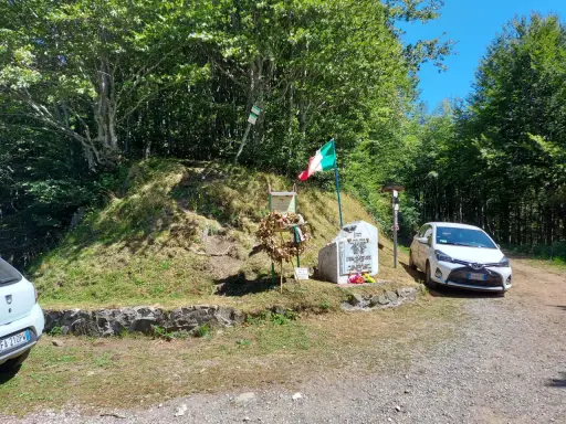 Monumento commemorativo con bandiera italiana e corona d'alloro al passo delle radici, circondato da alberi e con un'auto parcheggiata.