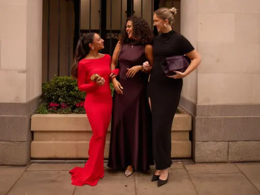 Three women in elegant gowns, discussing what not to wear to a wedding. One in red, one in plum, and one in black.