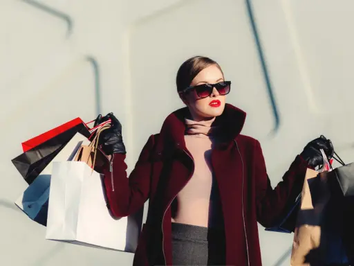 Donna con occhiali da sole e rossetto rosso, avvolta in un cappotto bordeaux, porta borse della spesa, simbolo di moda veloce.