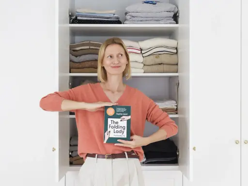 Kobieta z książką "The Folding Lady" pokazuje, jak składać koszulki, stojąc przed otwartą szafą pełną schludnie ułożonych ubrań.