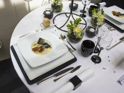 Une table de restaurant dressée avec élégance, un plat de poisson en vedette, des verres noirs et des fleurs.