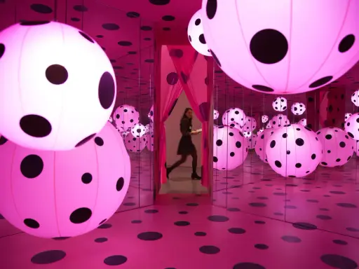 A woman walks through a mirrored room filled with pink polka-dotted spheres, a signature of Yayoi Kusama's immersive art.