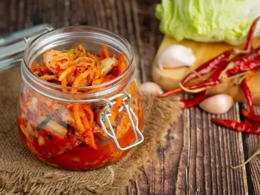 Słoik z kimchi na tle desek, obok kapusta, czosnek i papryczki chili.