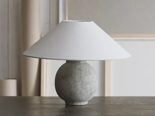 Bordslampa med rund, texturerad fot av lufttorkande lera, målad i grågröna toner. En vit lampskärm sitter ovanpå.