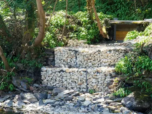 Estructura de muro de gavion con piedras blancas, rodeada de vegetación y rocas junto a un río.
