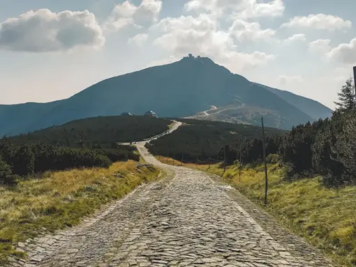 Kamienista droga prowadzi ku górze, gdzie jest Śnieżka. W oddali widać szczyt z budynkami i krętą ścieżkę.