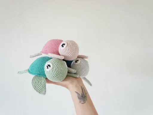 Una mano sostiene un trío de adorables tortugas a crochet: una verde menta, una rosa y una gris claro.