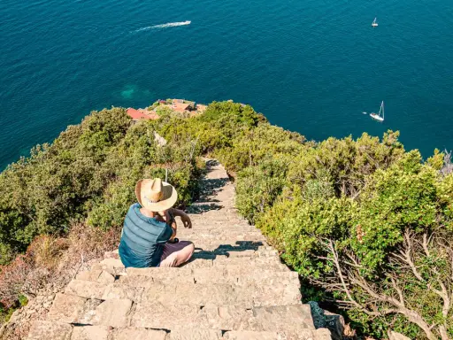 Un uomo con cappello di paglia seduto su una scalinata di pietra guarda il mare blu. Un sentiero scende verso il mare, suggerendo come arrivare a Monesteroli.
