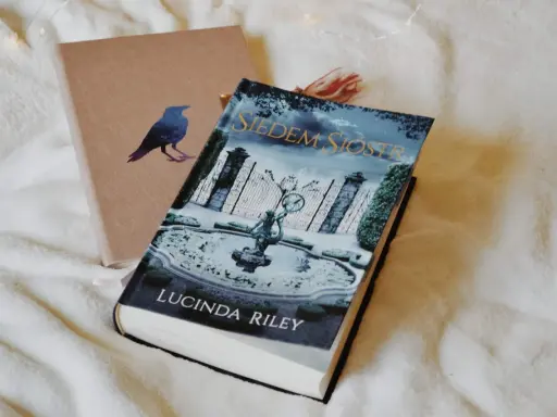 Okładka książki "Siedem Sióstr" Lucindy Riley na białym tle. Obok leży brązowy notes z wizerunkiem kruka.