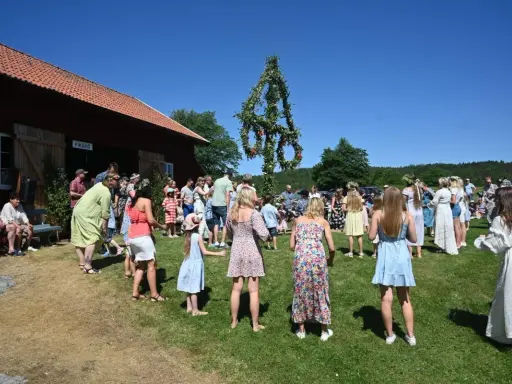 Folk dansar runt midsommarstången på midsommar afton. En vacker sommardag med klarblå himmel.
