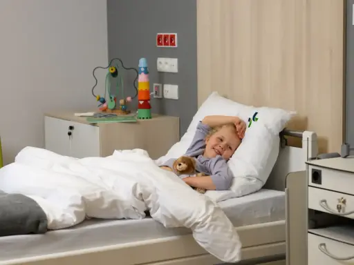 Uśmiechnięte dziecko w szpitalnym łóżku, przytulające misia. Obok zabawka edukacyjna i szafka.