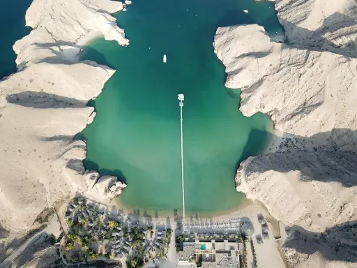 Vista aerea di un resort in Oman con bambini che giocano sulla spiaggia. Acque turchesi, montagne rocciose e un lungo pontile.