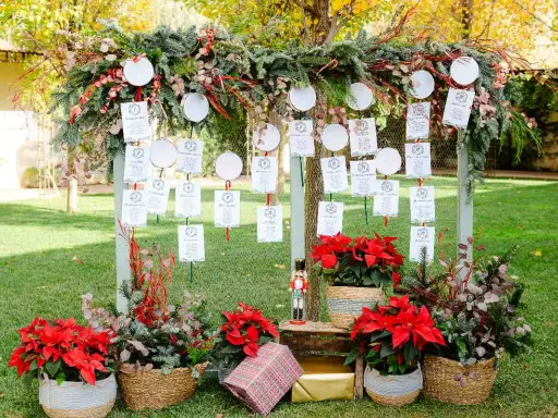 Decoración ceremonia civil con un arco de ramas y hojas, colgando tarjetas de invitados. Flores rojas y cestas adornan la base.