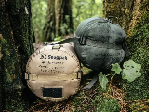 Dwa śpiwory Snugpak, jeden beżowy (komfort -7°C), drugi zielony (komfort 0°C), leżą na leśnym poszyciu.