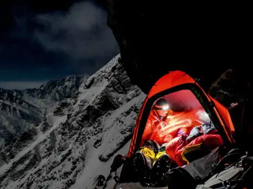 Nocny obóz na zboczu himalajskiego parbatu. W pomarańczowym namiocie rozświetlonym czołówką odpoczywa alpinista, otoczony surowym, ośnieżonym krajobrazem.