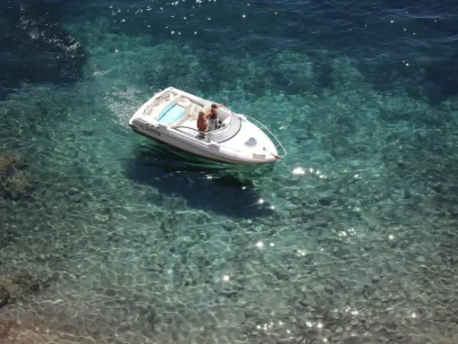 Deux personnes dans un bateau blanc, au moteur en panne, dérivent sur une eau turquoise et claire, révélant les fonds rocheux.