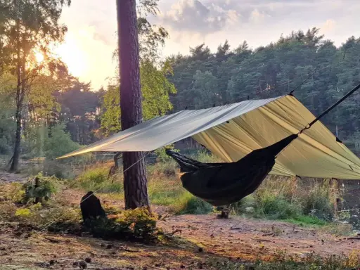 En hängmatta hänger under ett stort vindskydd presenning i en skog vid solnedgången.