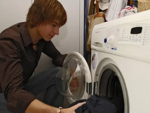 Junger Mann lädt Wäsche in die Waschmaschine. Bald werden die Sitzkissen frisch gewaschen sein.