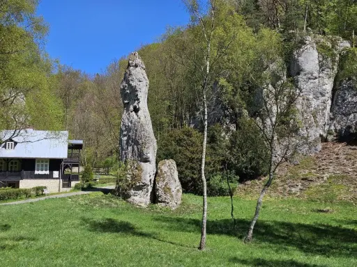 Imponująca skała Igła Deotymy góruje nad zieloną polaną, obok której stoi drewniany domek.