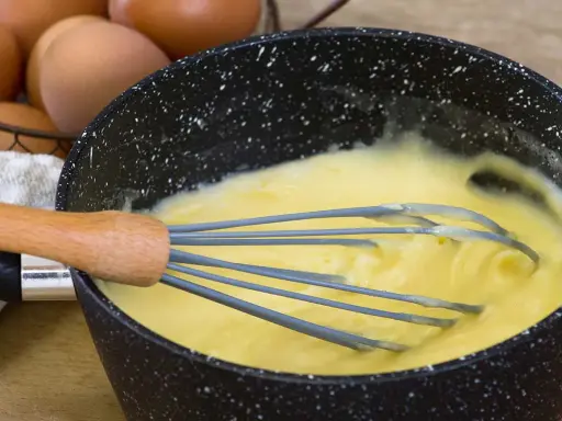 Un fouet mélange une crème pâtissière traditionnelle onctueuse dans une casserole. Des œufs sont visibles en arrière-plan.