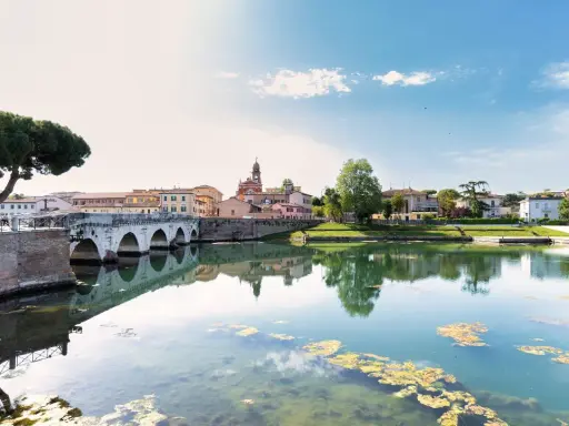 Ponte di Tiberio e il fiume Marecchia a Rimini a settembre, con riflessi sull'acqua e un cielo azzurro con nuvole bianche.