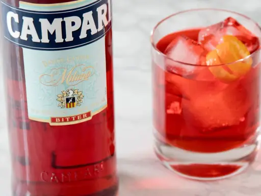 Butelka Campari i szklanka z czerwonym drinkiem, lodem i skórką pomarańczy. Campari to gorzki aperitif.