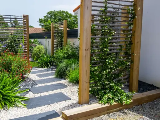 Un chemin de dalles grises serpente à travers un jardin luxuriant. Des treillages en bois servent de support à une plante brise vue grimpante, créant un espace intime.