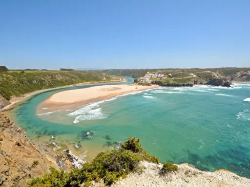 Atemberaubende Aussicht auf Praia de Odeceixe, wo ein Fluss auf den Ozean trifft. Grüne Hügel und ein weißes Dorf säumen die Küste.