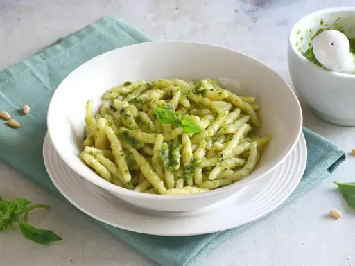 Un piatto di pasta con pesto fresco, guarnito con basilico e pinoli. Accanto, un mortaio con altro pesto.