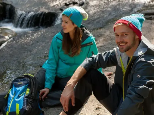 Para, co to jest trekking? Para w czapkach i kurtkach siedzi przy strumieniu, obok plecak.