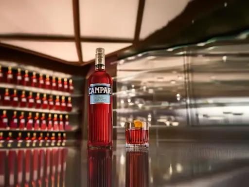 Eine Flasche Campari und ein Glas mit dem roten Aperitif stehen auf einer glänzenden Theke. Die Frage "wie schmeckt Campari?" wird durch den Anblick des ikonischen Getränks aufgeworfen.