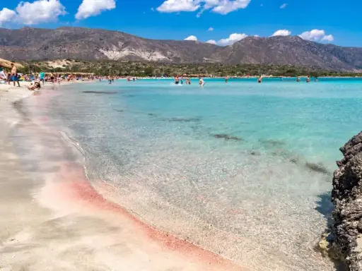 Spiaggia rosa di Creta con bambini che giocano nell'acqua cristallina e montagne sullo sfondo.