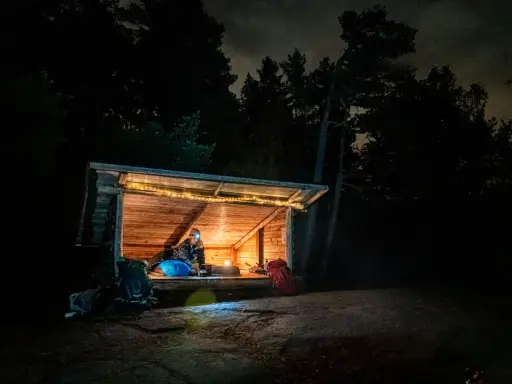 Person myser i ett upplyst vindskydd i skogen på natten. Ryggsäckar står utanför.