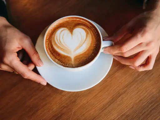 Eine Tasse Milchkaffee mit Herz-Latte-Art, gehalten von zwei Händen. Ein perfekter Start in den Tag, was ist Milchkaffee?